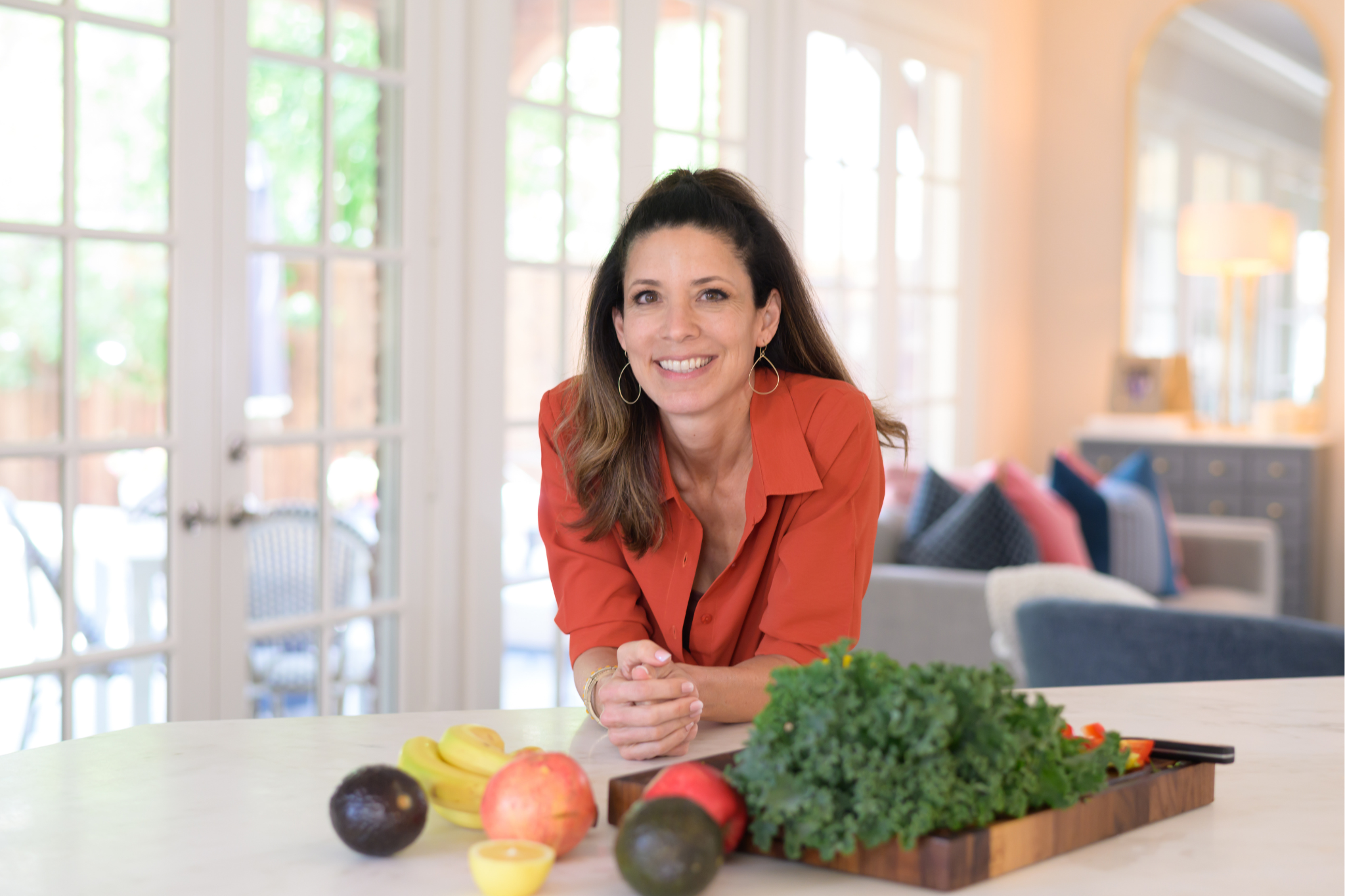 Kristi Phillips smiling, leaning on her kitchen counter beside fresh kale, fruit and vegetables